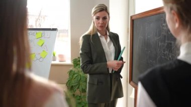 Confident math teacher explaining lesson standing at blackboard talking writing with chalk. Shooting over shoulders of curios teenage students listening tutor in classroom. Education and curiosity