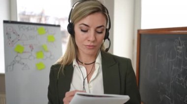 Video chat POV of positive confident teacher in headphones listening writing with pen. Professional Caucasian beautiful woman teaching online on lockdown standing in classroom with whiteboard