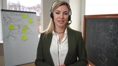 Portrait of confident woman in headphones talking looking at camera standing in classroom with whiteboard and chalkboard. Professional psychologist teaching online. Blogging and convenience concept
