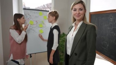 Happy psychology teacher crossing hands looking at camera smiling with teen students talking at whiteboard at background. Confident elegant Caucasian woman posing in classroom with schoolgirls