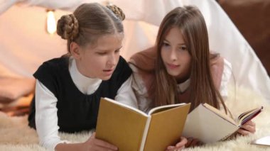 Intelligent curios teenage girls reading books talking lying indoors in living room with tent. Happy smart teen Caucasian friends discussing literature enjoying hobby. Education and friendship