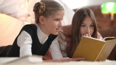 Excited absorbed teenage girls reading book talking lying indoors at tent in living room. Portrait of curios concentrated Caucasian teen friends enjoying hobby on weekend chatting