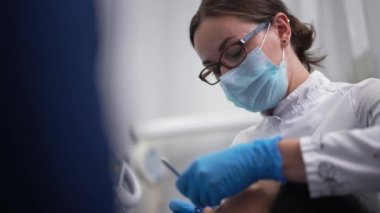 Portrait of intelligent confident female dentist in eyeglasses and face mask examining patient with dental mirror in slow motion. Caucasian doctor helping woman with toothache in hospital indoors