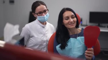 Portrait of satisfied patient with mirror admiring result as blurred dentist smiling at background. Happy Caucasian woman with professional doctor in hospital indoors. Slow motion