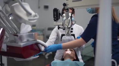 Dental microscope with professional doctor examining patient and assistant moving tools closer in slow motion. High-quality equipment and expert dentist treating Caucasian woman in hospital