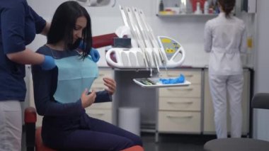 Side view woman sitting in dental chair as assistant putting on dental napkin and dentist walking from background. Caucasian patient on doctor visit in hospital indoors with doctor and nurse
