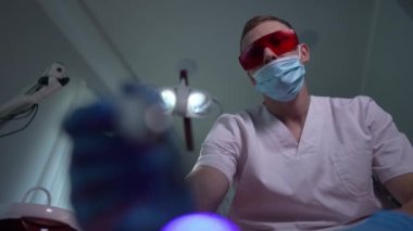 Patient point of view of Caucasian dentist in uniform and protective eyeglasses using flashlight examining oral cavity. Bottom angle view portrait of concentrated professional doctor in hospital