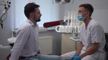Side view positive male patient and dentist talking in slow motion sitting in hospital indoors. Medium shot portrait of confident professional doctor and Caucasian man discussing pain treatment