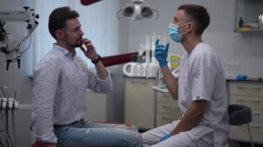 Side view dentist asking questions talking with Caucasian man sitting in dental chair. Portrait of professional confident doctor consulting patient in medical clinic indoors. Slow motion