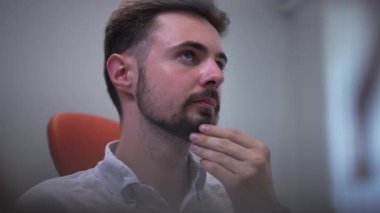 Close-up thoughtful handsome man rubbing beard sitting in dental chair waiting. Portrait of confident Caucasian male patient in medical clinic on appointment visit. Slow motion