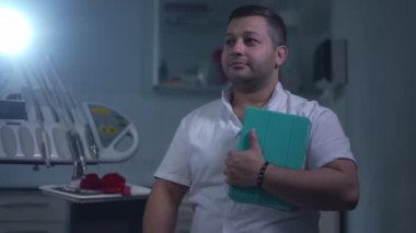 Medium shot of confident Middle Eastern doctor with digital tablet looking away turning looking at camera smiling. Positive professional dentist posing in slow motion at background of dental light