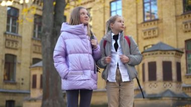 Two sad Caucasian teen schoolgirls standing on schoolyard talking with dissatisfied facial expressions. Adolescent teenage classmates not willing go to school. Lifestyle and education concept