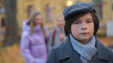 Portrait of Caucasian bullied schoolboy looking at camera as blurred classmates laughing at background. Sad overwhelmed depressed cute boy posing on schoolyard on autumn day. Bullying concept