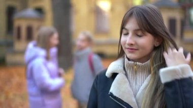Self-assured adolescent schoolgirl posing on schoolyard with blurred classmates at background. Portrait of confident Caucasian teen student looking at camera smiling touching hair. Adolescence