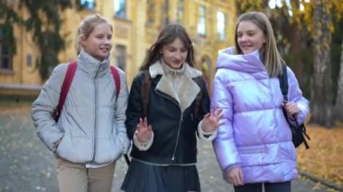 Tracking shot of three positive female teen classmates walking after classes gossiping. Beautiful happy Caucasian schoolgirls talking strolling outdoors on schoolyard. Live camera follows students