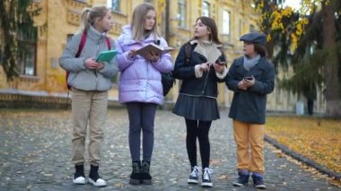 Wide shot portrait of nerd intelligent teenage classmates talking discussing studies on schoolyard. Happy confident Caucasian girls and boy with tablet phones and notebook outdoors