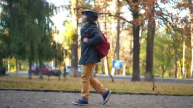 Side view curios cute boy with rucksack walking on autumn city street looking around. Wide shot of confident smart Caucasian schoolboy with backpack strolling outdoors after classes on autumn day