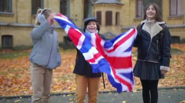 Joyful confident school children shaking British flag laughing looking at camera. Positive smart boy and girls posing on schoolyard outdoors on autumn day having fun. Patriotism and education