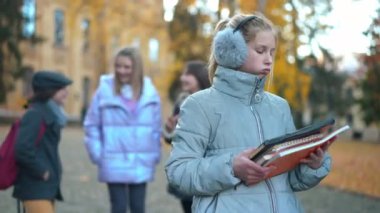 Exhausted overwhelmed teen girl standing with homework on the right as blurred classmates talking at background. Portrait of tired Caucasian schoolgirl looking away thinking. Education load