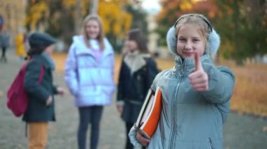 Smart confident teen schoolgirl gesturing thumb up smiling looking at camera standing outdoors with blurred classmates at background. Portrait of happy intelligent Caucasian girl posing on schoolyard