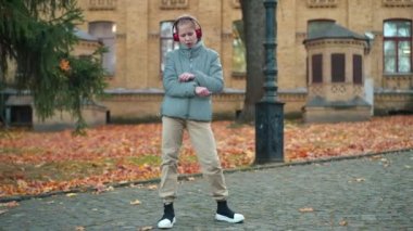 Wide shot confident teenage girl in headphones dancing listening to music on schoolyard. Live camera zoom in to Caucasian teen schoolgirl having fun enjoying hobby on break outdoors