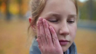Headshot of sad frustrated cute teen schoolgirl touching cheek and looking at camera with depressed facial expression. Close-up portrait of bullied Caucasian teenage girl posing in schoolyard