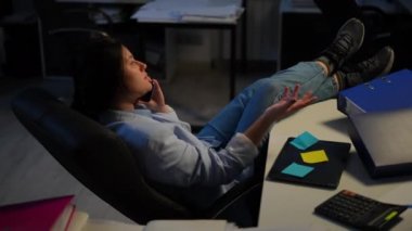 Young woman with feet on table sitting in office talking on phone juggling tangerine. Wide shot portrait of confident Caucasian female consultant indoors. Planning and business concept
