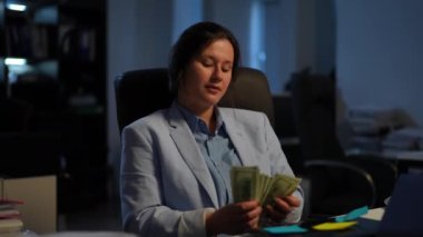 Portrait of satisfied rich manager counting cash sitting at table in office indoors. Happy confident Caucasian young wealthy woman at workplace with money. Success and achievement concept