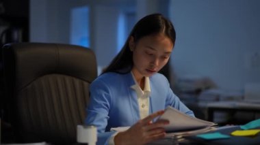 Concentrated beautiful Asian woman thinking analyzing paperwork in office in the evening. Portrait of intelligent young manager overworking in darkness indoors. Business and analysis concept