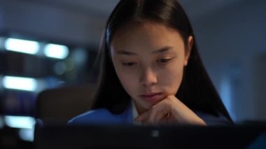 Close-up front view of young Asian woman thinking surfing Internet on laptop in darkness. Portrait of smart confident beautiful employee analyzing project messaging online in office indoors