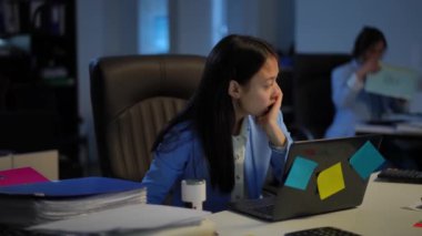 Troubled tired Asian woman sighing surfing Internet on laptop leaning back on chair as blurred Caucasian colleague at background leaving with document folders. Portrait of fatigue employee in office
