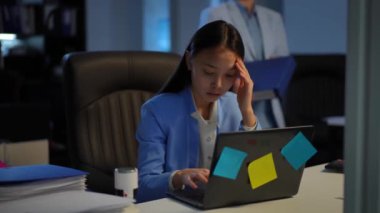 Asian woman working in office as Caucasian colleague passing bringing paperwork. Portrait of sad stressed young employee sitting with laptop indoors looking at camera sighing. Multitasking concept