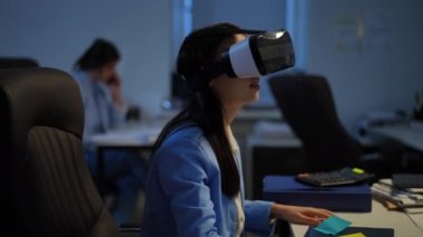 Side view absorbed excited Asian woman sitting in office gaming online in VR headset. Concentrated slim young employee enjoying break at workplace indoors. Augmented reality concept