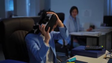 Positive Asian woman passing VR headset to Caucasian colleague at background smiling. Portrait of relaxed carefree young employees having fun at workplace in the evening. Augmented reality concept