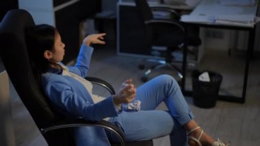 Side view bored Asian young woman throwing paper in trash bin. Slim female employee sitting on chair wasting time procrastinating in office indoors. Lifestyle and boredom concept