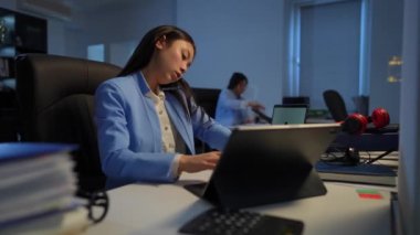 Busy intelligent Asian woman talking on phone messaging online on laptop sitting in office with Caucasian colleague working at background. Concentrated smart confident employee at workplace indoors