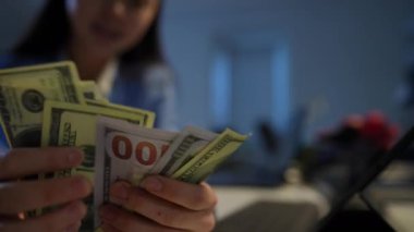 Close-up cash dollars in hands of unrecognizable Asian woman counting income. Happy satisfied businesswoman in suit sitting in office indoors. Success and business concept