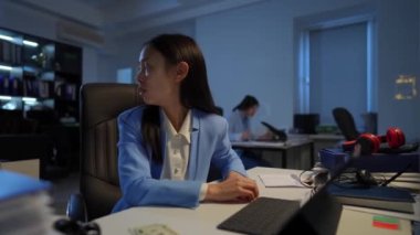 Stressed overburdened Asian woman having panic attack as angry Caucasian colleague arguing at background. Portrait of anxious worried employee breathing in paper envelope sitting in office
