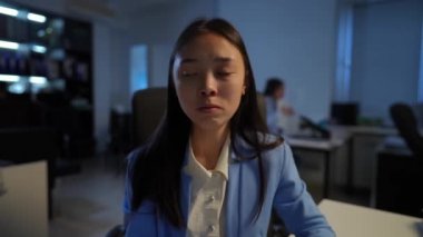 Asian young woman with symptoms of panic attack breathing in paper envelope sitting in office with blurred Caucasian coworker at background. Portrait of troubled worried employee at workplace