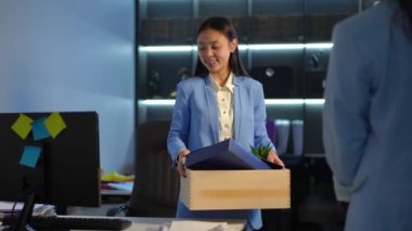 Happy new employee standing with box at workplace as manager greeting Asian woman in office leaving. Portrait of satisfied confident beautiful woman indoors looking around smiling
