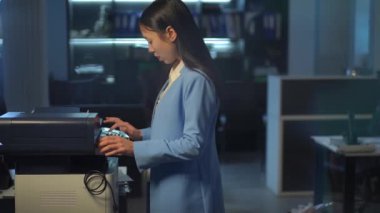 Busy Asian young woman in elegant suit walking to printer in office printing document walking to table. Confident manager working indoors in the evening. Business and technology