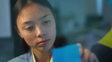 Headshot portrait of focused Asian young woman writing with pen on adhesive note on glass. Close-up of concentrated intelligent smart manager planning schedule in office indoors