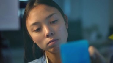 Front view close-up portrait of Asian woman writing with pen on adhesive notes on glass walking away leaving. Headshot of intelligent confident manager planning schedule in office