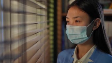 Portrait of young Asian charming woman in Covid-19 face mask looking out the window and looking at camera standing in office. Confident female employee posing indoors on coronavirus pandemic