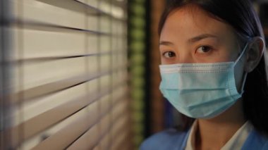 Headshot portrait of confident Asian young woman in coronavirus face mask posing in office at window with shades. Close-up face of intelligent female employee looking at camera standing indoors