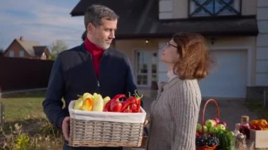 Confident adult couple of Caucasian farmers posing with basket of belly pepper outdoors. Portrait of positive husband and wife looking at camera smiling selling vegetables on front yard