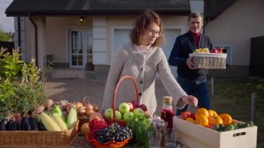 Positive Caucasian woman standing at table on front yard with fruits and vegetables as man bringing box of belly pepper talking. Adult couple of farmers selling healthful organic products outdoors