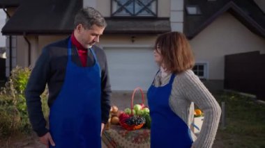 Medium shot of smiling adult Caucasian man and woman in blue aprons looking at camera smiling selling organic products. Confident couple of gardeners posing at table with fruits and vegetables