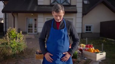 Portrait of confident male farmer in blue apron crossing hands looking at camera standing on front yard. Handsome man posing at table with organic fruits and vegetables for sale. Lifestyle and farming