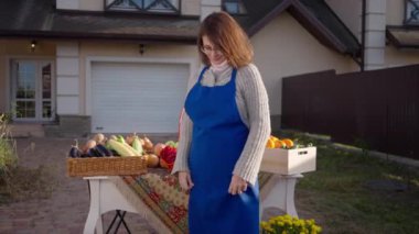 Confident female farmer in eyeglasses and blue apron crossing hands looking at camera standing on front yard selling fruits and vegetables. Positive Caucasian woman posing on sunny autumn day outdoors